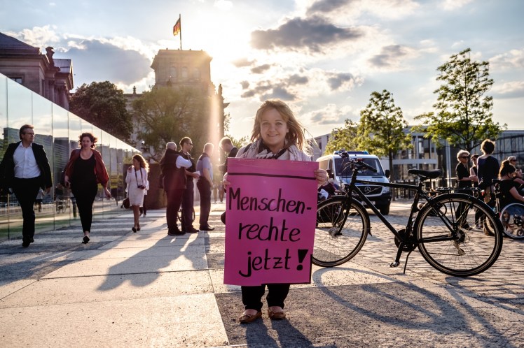 Foto: Kleinwüchsige blonde Frau lächelt in die Kamera und hält ein pinkes Schild in der Hand, auf dem steht: Menschenrechte jetzt!