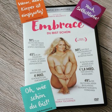 Foto: Die DVD-Hülle der Doku EMBRACE ist zu sehen. Darauf liegen drei farbige Sticker mit jeweils dem Aufdruck: "Wow, dein Körper ist einzigartig!", "Yeah, Selbstliebe!" und "Oh, wie schön du bist!"