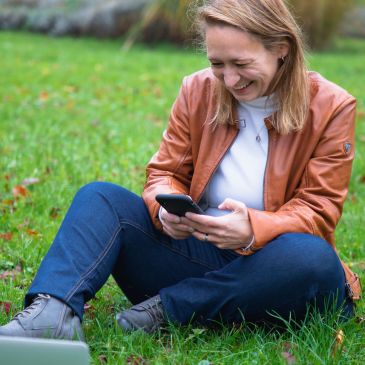 Nadine sitzt lachend auf einer Wiese und schaut auf ihr Smartphone