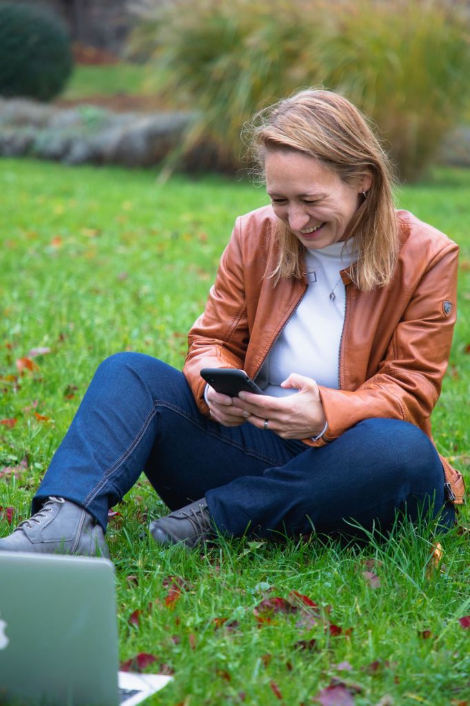 Nadine sitzt lachend auf einer Wiese und schaut auf ihr Smartphone